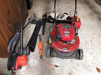 Overall view of lot showing Craftsman red mower, Toro blower vac with hose and collection bag, and Black & Decker orange edging tool and electric trimmers on concrete floor.