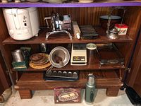 Shelf with Sunbeam fryer on left, vintage meat slicer center top, cornbread pans on right top shelf, meat grinder bottom left, vintage percolator, baking dish, and thermos