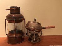 Photo showing both the Dutch copper seaman's lantern and the Thai brass fondue set placed side by side on a wooden surface.