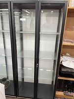 Three black-framed glass display cabinets with locking doors shown side by side. Each has four glass shelves inside and keys for the lock.