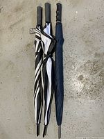 Photo of three large umbrellas laid flat on concrete floor. One umbrella is solid blue and two umbrellas have black and white stripes. All are folded and have hooked black handles.