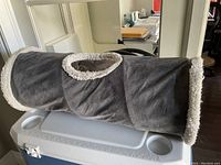 Image of a soft grey cat play tube with white fluffy trim, showing side circular openings and the tube placed on a cooler.