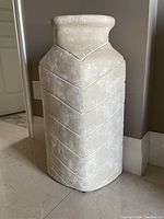 Tall beige ceramic vase with geometric embossed pattern on floor next to wall.