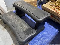 Photo of black plastic two-step hot tub or pool steps, showing curved edges and textured surface, some dust visible, sitting on a blue tarp with wood and cords in background.