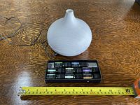 Photo showing a white round-shaped electric diffuser with wood-grain texture and narrow top opening. Below it is a black box containing 8 visible small bottles of essential oils with colored labels arranged neatly in two rows. A measuring tape placed in front shows approximately 8 inches wide diffuser.