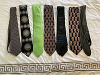 Seven men's neckties shown laid flat in a row on a white patterned cloth background.