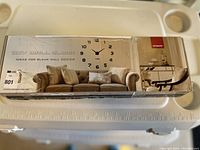 Photo of the DIY wall clock kit packaging showing a modern clock design with large black numbers, clock mechanism, and installation image above a sofa.