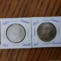 Front side of 1967 and 1968D Kennedy half dollar coins in white holders showing profile of JFK with inscriptions and dates.