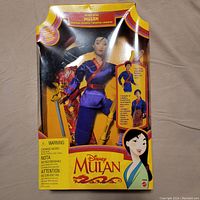 Front view of the Disney Mulan Secret Hero doll in original packaging showing doll dressed in purple outfit with sword and scroll included.