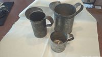 Three antique tin items including two pitchers and a cup displayed on a white cloth surface.