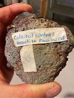 Hand-held front view showing specimen and handwritten label 'Calcite coated hematite Faraday, ON'