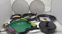 Photo of various frying pans and mesh splatter guards arranged showing handles, lid, and pan surfaces.