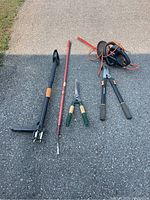All garden tools displayed on pavement including a Fiskars weeder, hedge shears, pruners, and electric hedge trimmer.