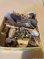 Top view of box containing an assortment of rock and mineral specimens, some with visible labels including Tigre, Africa and a note describing bright pyrite and quartz cluster.