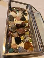 Top view shot showing various colorful polished and raw rock, mineral, and crystal specimens inside a rectangular glass and metal display box.