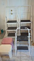 Photo showing two white metal step ladders with three black treads each, a small black metal two-step stool, and two plastic step stools (pink and cream) arranged on a tiled floor in front of a closed door.