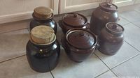 Photo showing all six vintage ceramic crock jars arranged on tile floor, mix of wooden and ceramic lids visible.