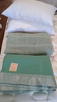 Photo of stacked pillows and blankets on mattress showing green textured blanket, green wool blanket with satin edges, and two white pillows.