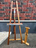 Wooden stand-up art easel and wooden plein air canvas carrier placed outdoors against a brick wall, showing overall condition and design of both items.