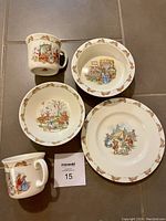 Full set of Bunnykins dishes including two mugs, one dinner plate, one rimmed bowl, and one bowl arranged on tile floor.