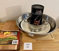 Lot contents shown with large stainless steel bowl, white ceramic serving bowl, small metal colander inside, and black cylindrical glass decanter placed in colander. Cedar grilling planks and wooden cutting board shown nearby.