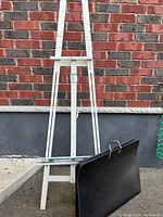 Photo showing a white wooden easel and a large black portfolio case standing against an outdoor brick and concrete wall.