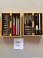 Two yellow plastic carrying cases open to display Exacto precision knives, various blades, multi-tool, and handles inside, arranged neatly and fully visible.