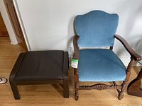 Antique wooden chair with blue velvet upholstery and IKEA Poang footstool with black leather or leather-like seat shown side by side on wooden floor against white wall.