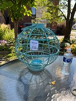 Photo showing a large spherical turquoise metal garden lantern candle holder placed outdoors on a glass table, next to a water bottle for scale.