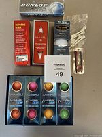 Photo showing multiple boxed golf balls from Dunlop, Maxfli, Top-Flite, and Noodle Ice series, plus golf tees and a ball marker tool.