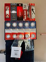 Photo showing golf balls in original boxes stacked in two rows with two boxes of Noodle brand showing 5 balls each in a lower row. Two separate plastic bags containing white golf tees and a black towel with clip visible underneath.