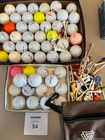 Two boxes filled with assorted used golf balls in various colors, a pile of assorted golf tees in multiple colors and materials, and a small brown zippered pouch.