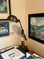 Side view of the metal industrial style lamp positioned on a corner desk with framed artwork in the background, showing its general size and shape.