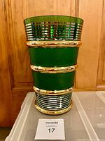 Photo of a tall emerald green and gold glass vase with silver horizontal stripes, showing the full front view of the vase on a flat surface with a wooden background.