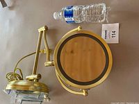 Full view of the brass-colored wall-mounted magnifying mirror with adjustable arm extended, accompanied by a water bottle for scale.