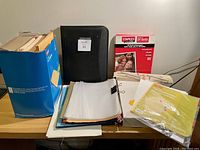 Photo shows various office supplies on a table including envelopes, a black binder, unopened Staples printer paper, clear plastic sleeves, file folders, and loose sheets.