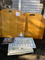 Two large wooden boards with carry handles standing upright behind two plastic paint palettes and a water bottle on a bench. A book titled 'Direct Water Colour' is positioned leaning against the palettes.