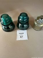 Three power line insulators displayed side by side on a beige surface, two teal green and one clear.