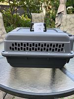 Front view of rectangular plastic pet carrier with ventilation slots and handle on top, placed on a glass table with garden chairs in the background.