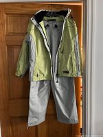 Full view of the women's rain suit showing the green and gray rain jacket and gray rain pants hung up together.