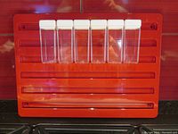 Front view of red plastic modular wall-mounted spice rack board measuring 16" x 11.5" with 6 clear plastic spice jars fitted in grooves, each having white lids.