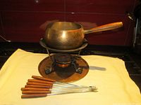 Copper fondue pot with wooden handle set on stand with copper underplate, burner and seven teak handled fondue forks laid in front on yellow cloth.