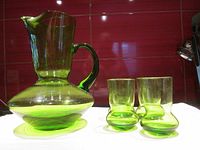 Green glass pitcher with wide base and curved handle alongside four matching glasses with rounded bottoms, photographed against a red tiled background.