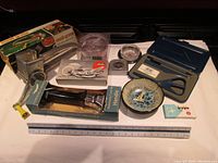 Photo showing collection of kitchen tools including the moulin-grater in box, scissors, glass bowl with screws, and measuring tape.