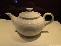 White teapot on a light cloth background, showing full side view including spout, handle, and lid with knob