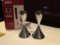 Pair of polished metal hourglass-shaped candlestick holders with box in background showing product image and branding.