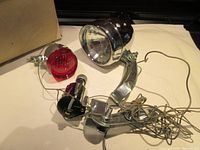 Two bike lights with wiring, red light attached to black generator clamp, and front headlight with chrome bracket and wire.