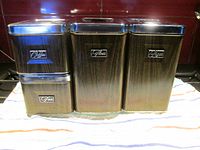 Set of four vintage metal kitchen canisters with chrome lids and faux wood grain finish, labeled Coffee, Flour, Sugar, and Tea, displayed on a cloth surface against dark background.