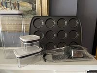 View of three clear rectangular OXO plastic containers with lids next to a dark non-stick muffin top pan with twelve circular molds.