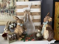 Photo showing three coir bunnies of varying sizes, a bunny-shaped striped wooden wall hanging with a bow and tag, a bag of small carrot decorative pieces, a box containing glass bunny table scatters, and other small decor elements.